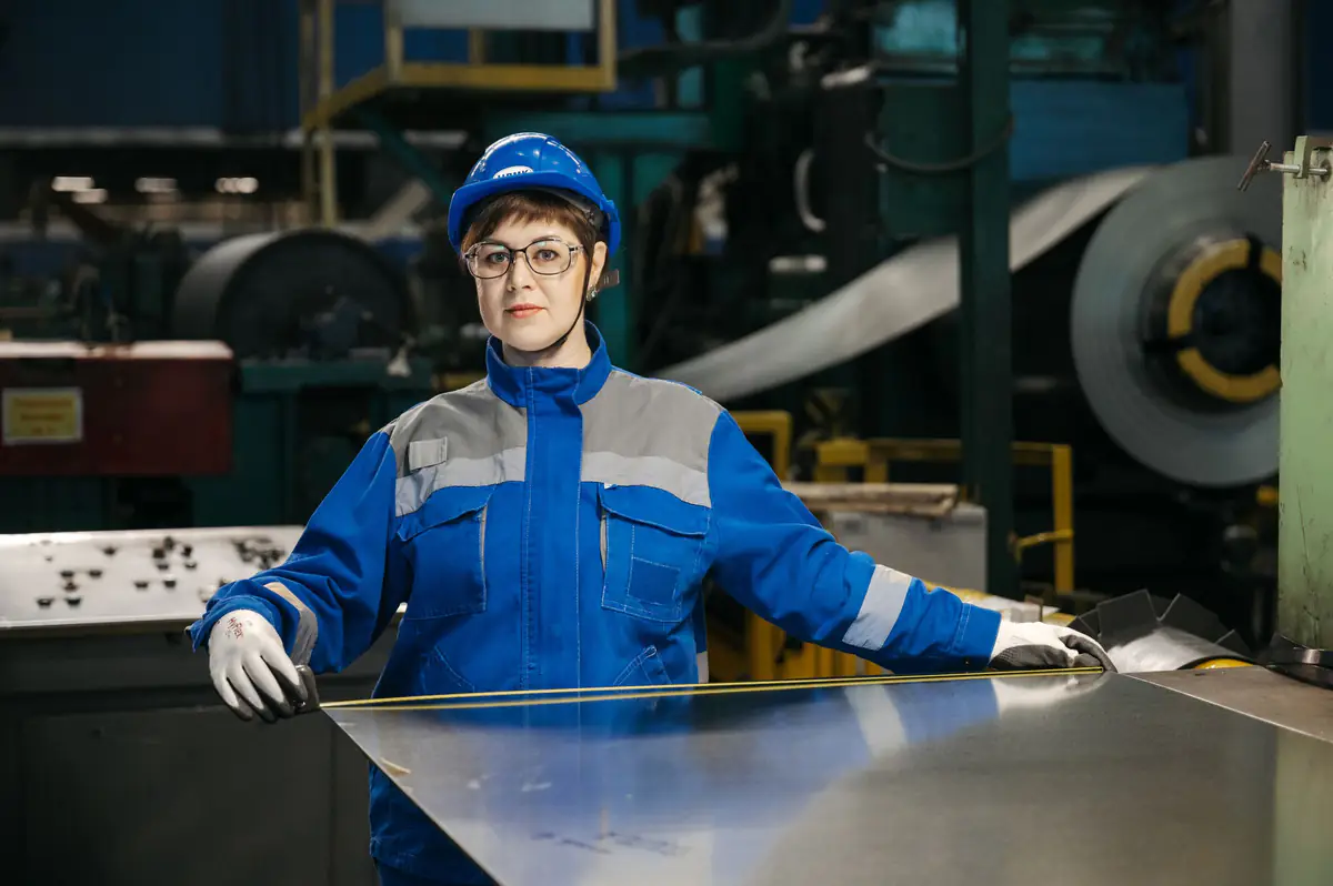 Profissional com equipamentos de proteção manuseando chapa metálica diante de bobina em ambiente industrial